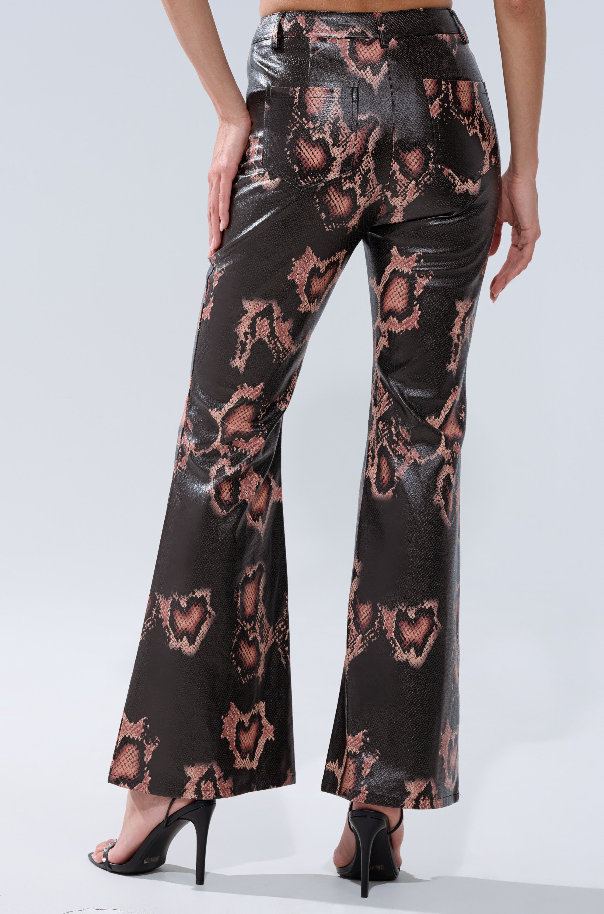 A person shown from the back wears high-heeled sandals and the COLD BLOODED FAUX LEATHER PANTS, featuring a brown snakeskin print with pink and black snake pattern.