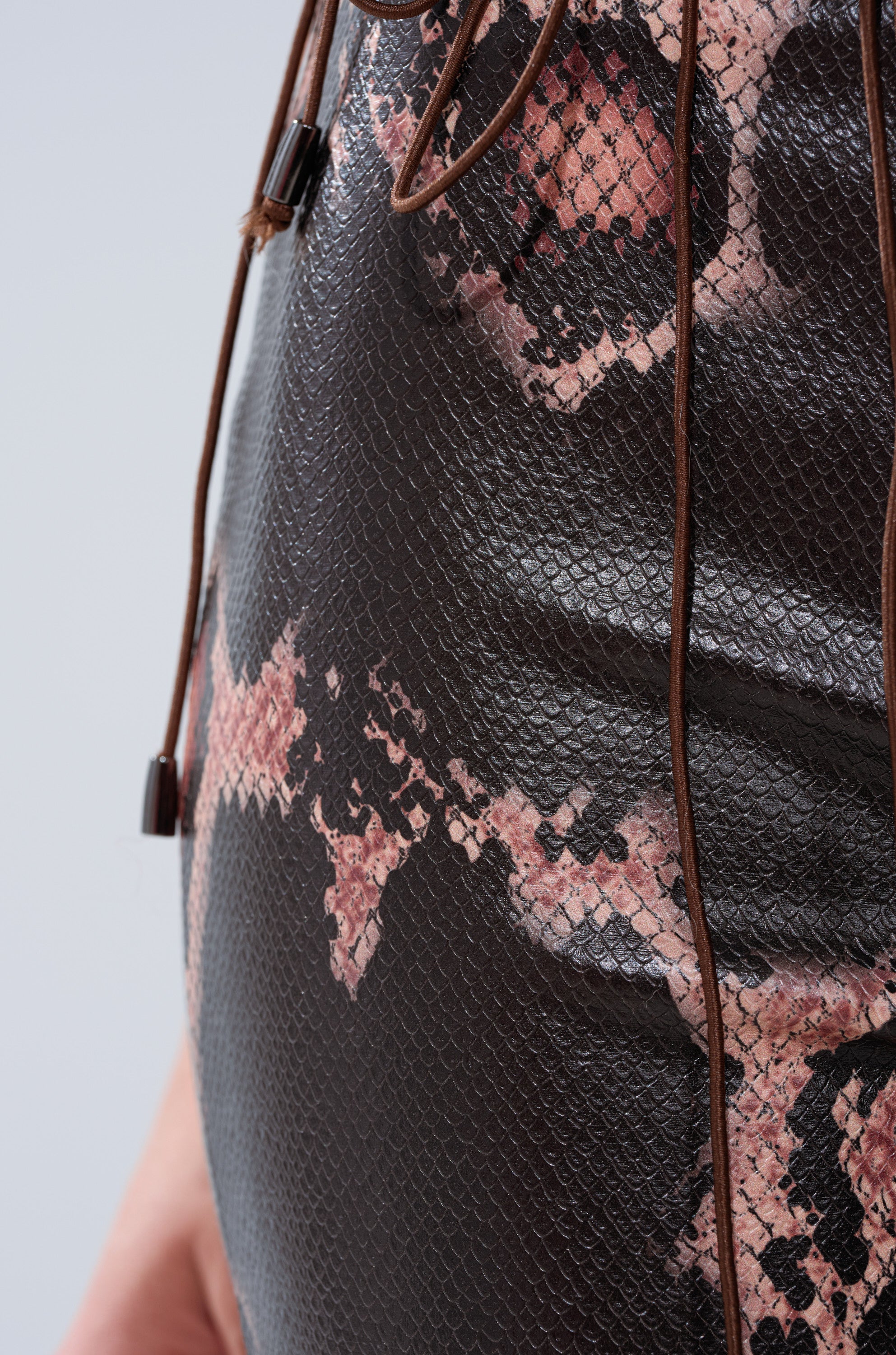 A close-up of the COLD BLOODED FAUX LEATHER PANTS featuring high-rise flared legs, shiny textured faux leather with a pink and black snakeskin pattern, and brown drawstring cords.