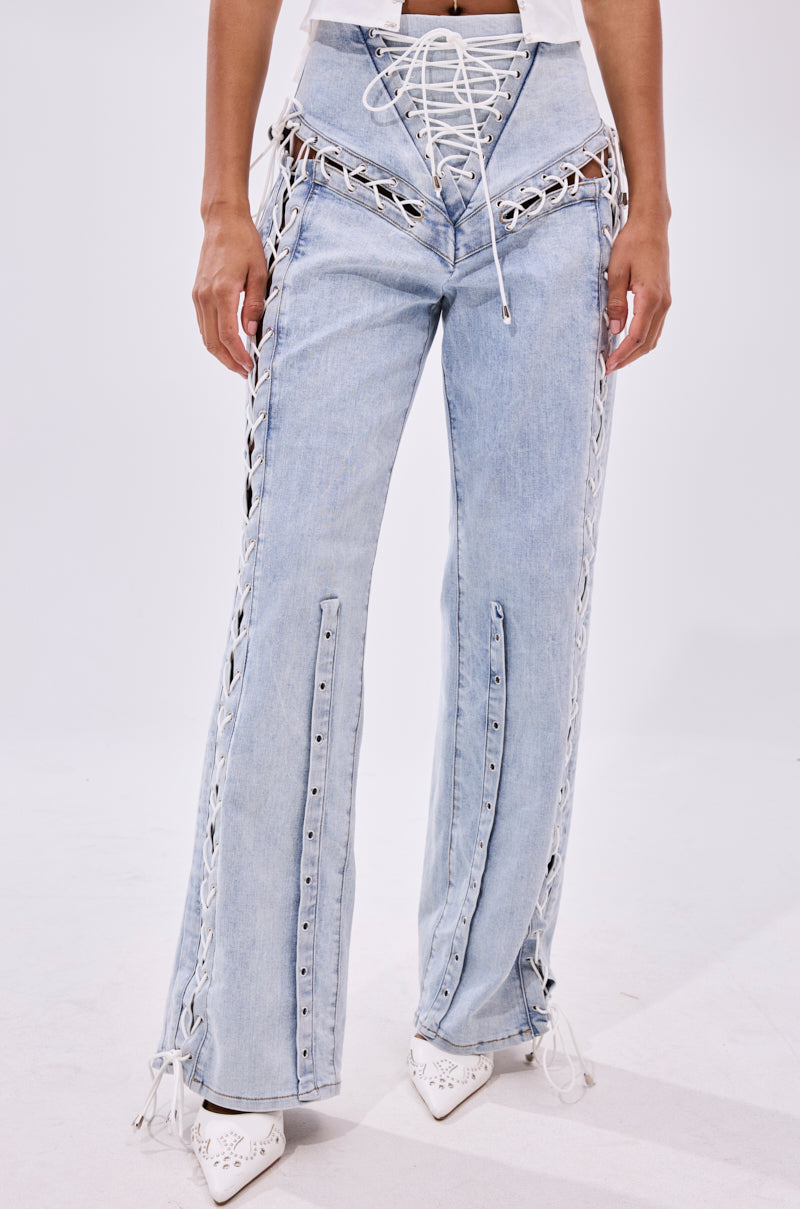 A person models the FEELING BLESSED LACE UP DENIM, light blue high-waisted jeans with white laces along the sides and front, styled with white pointed-toe shoes. The photo is cropped from shoulders down, highlighting the wide leg silhouette.