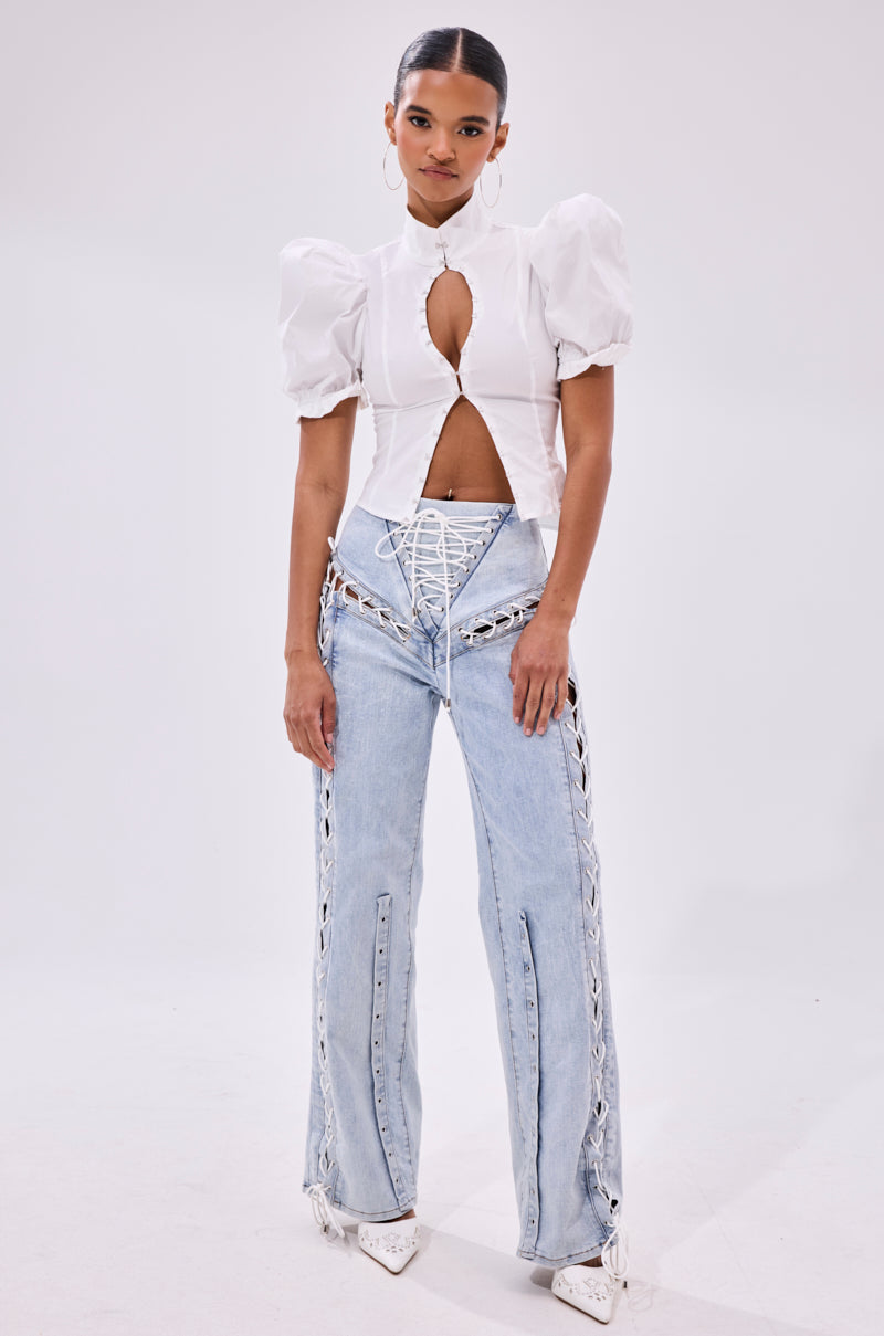 A woman poses against a plain background in a white puff-sleeve keyhole top and FEELING BLESSED LACE UP DENIM, styled with white pointed-toe heels and hoop earrings.