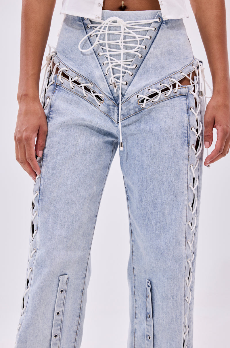 Model wears the FEELING BLESSED LACE UP DENIM in light blue, featuring white lace-up details, stylish cut-out sections, and a high waisted wide leg fit. Only the torso and legs are visible.