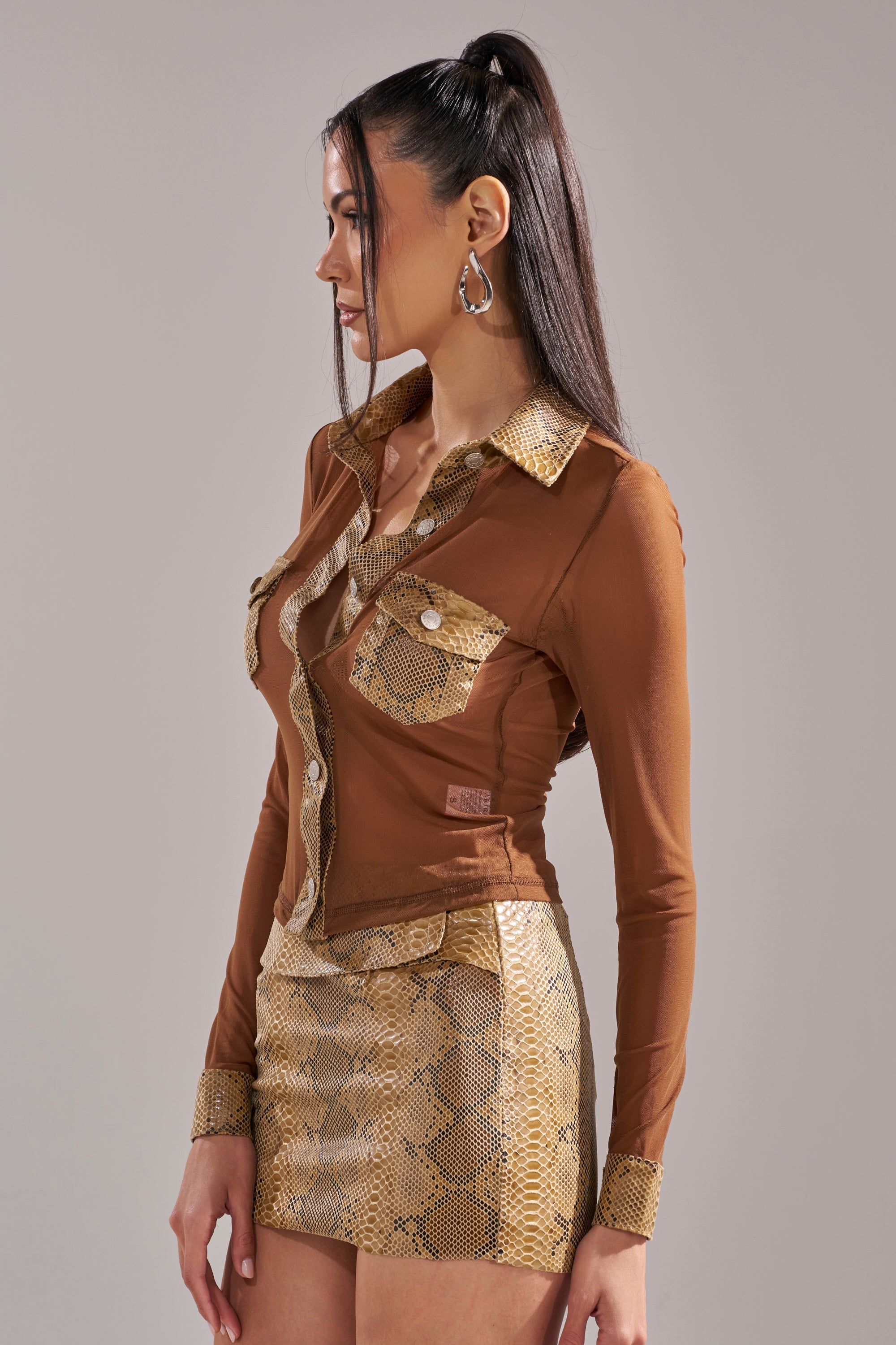 A woman with long dark hair in a ponytail models the FEELING SOME TYPE OF WAY FAUX LEATHER SNAKE BUTTON DOWN, a chic brown shirt with snakeskin-patterned collar, cuffs, and pockets, paired with a matching mini skirt against a plain background. 3