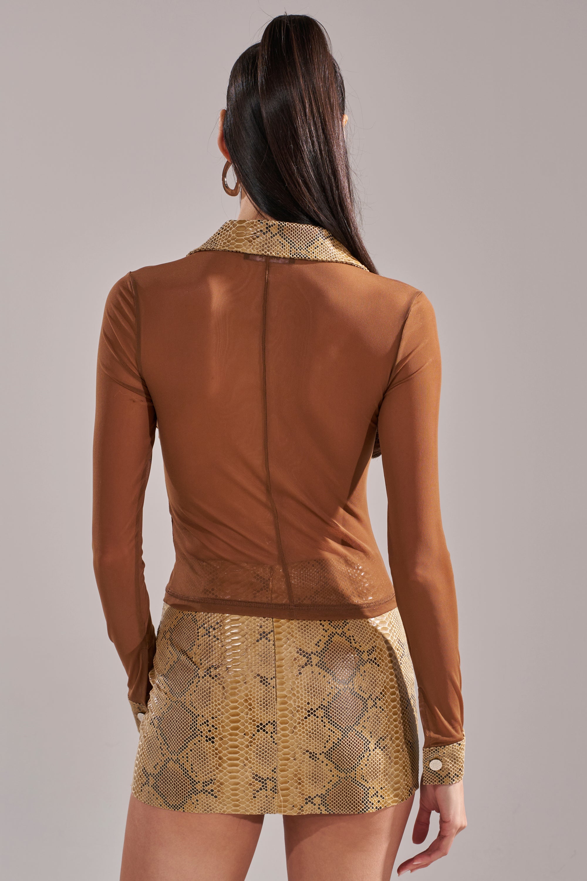 A woman with long brown hair in a high ponytail stands with her back to the camera, wearing the FEELING SOME TYPE OF WAY FAUX LEATHER SNAKE BUTTON DOWN and a high-waisted yellow snakeskin-print skirt. 4