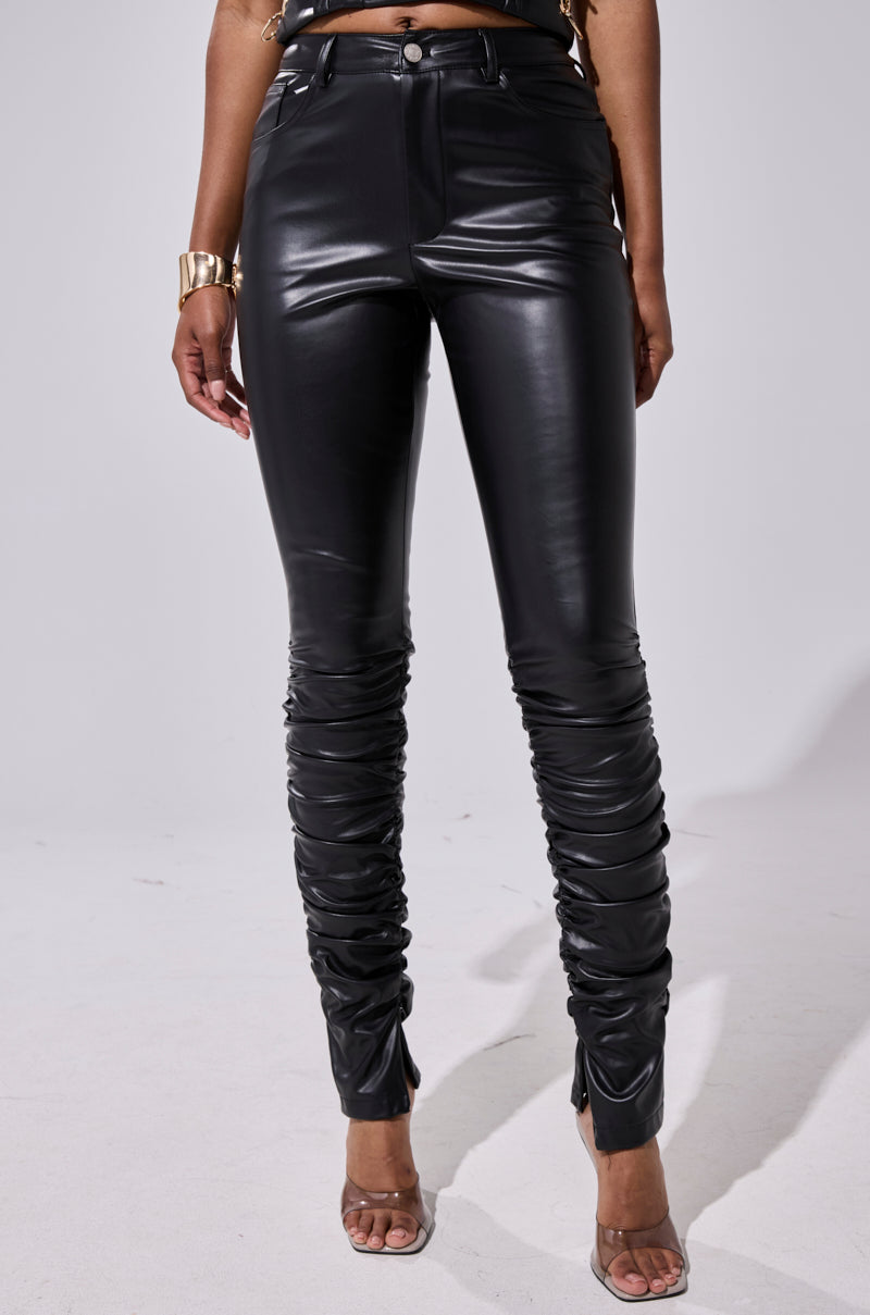 A person models the BADDIE STACKED FAUX LEATHER PANT with ruched details, paired with clear high-heeled sandals and a chunky gold bracelet. Their arms rest at their sides against a plain, light background. 2