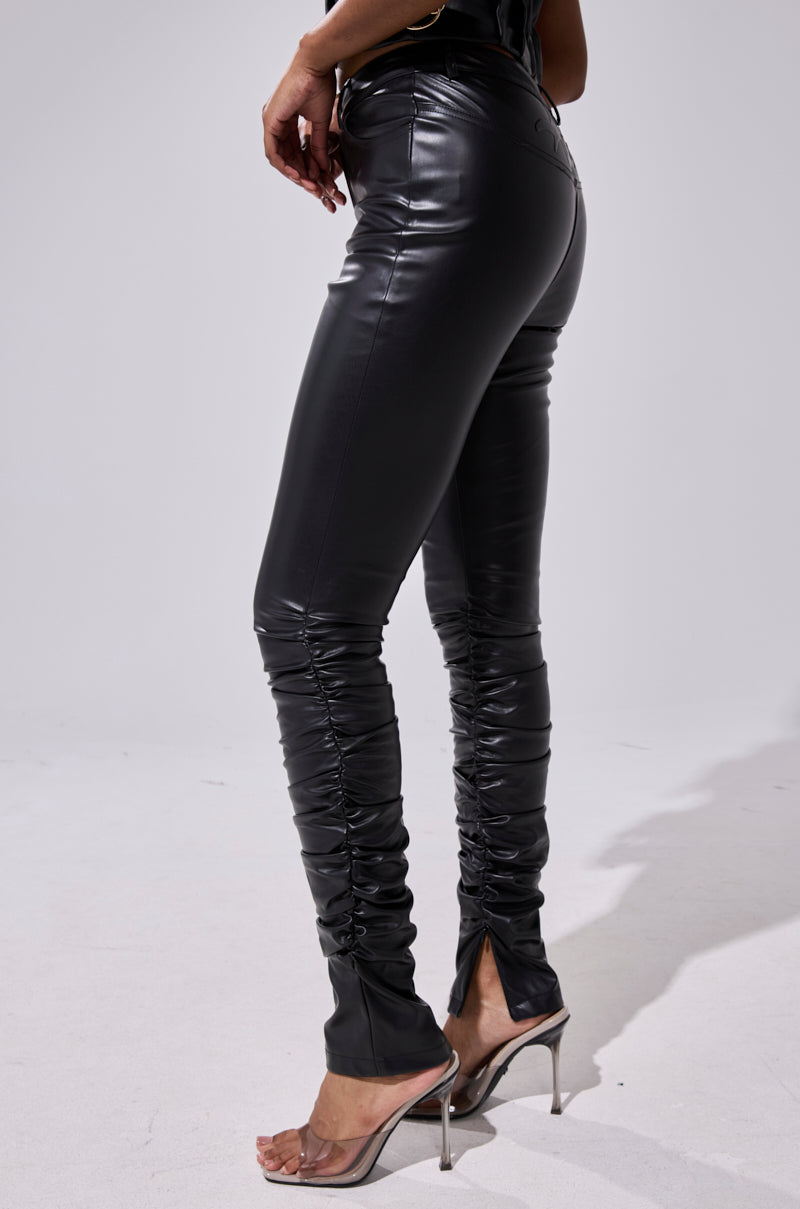 Someone stylishly stands on a light surface wearing the BADDIE STACKED FAUX LEATHER PANT in black, featuring ruched details and back ankle slits, paired with clear high-heeled sandals. 3
