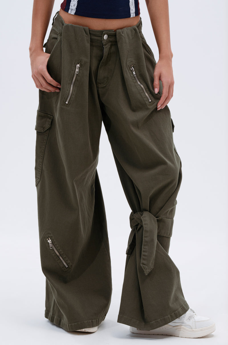 Wearing the COOL GIRL ERA TROUSER in olive green, featuring a wide-leg silhouette, multiple zippers, and large front pockets—one leg tied at the ankle—styled with a navy top and white sneakers.