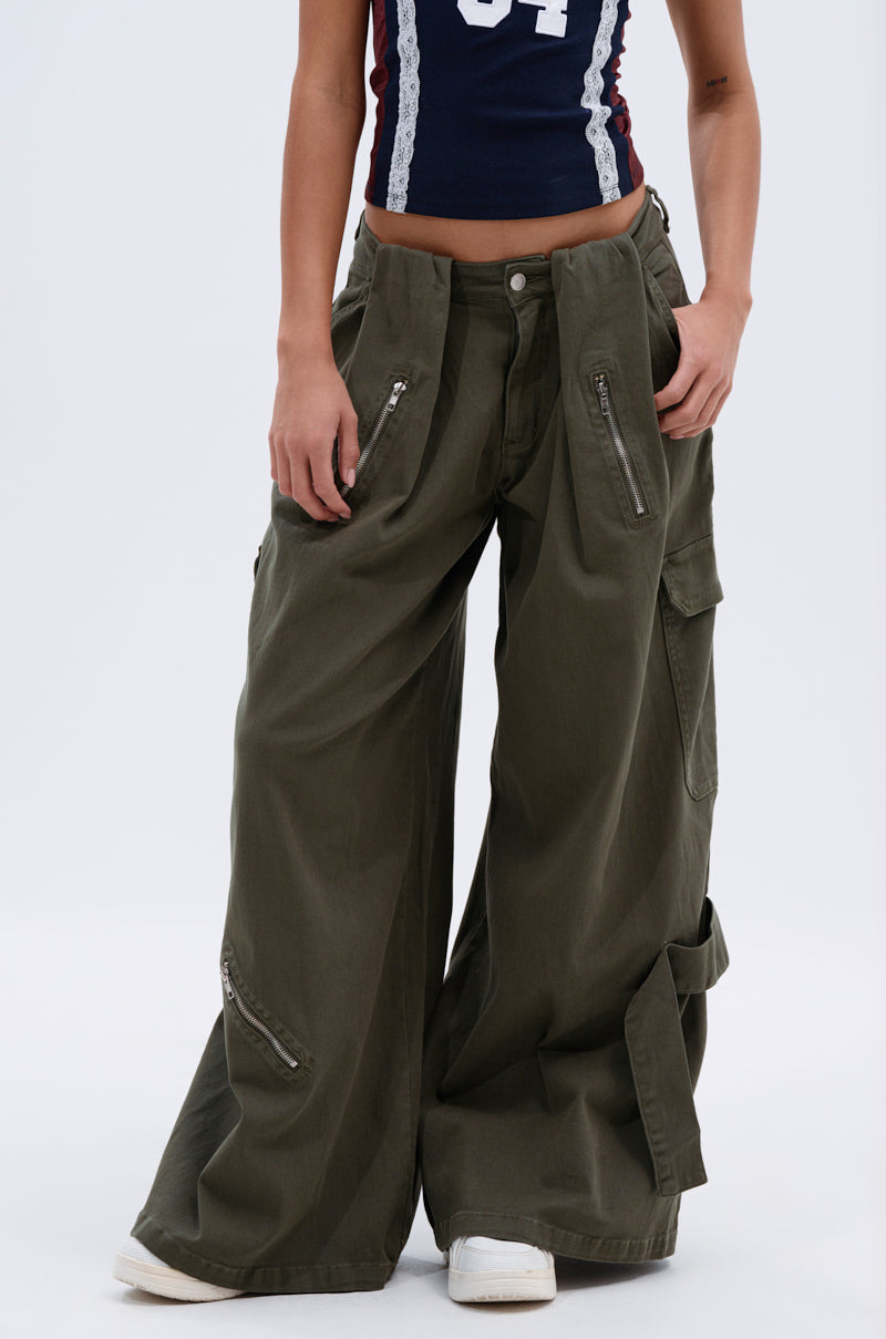 A person models the COOL GIRL ERA TROUSER in olive green, featuring wide-leg silhouette and multiple zipper pockets, styled with white sneakers and a sleeveless sporty top. The image is cropped at the shoulders and ankles.