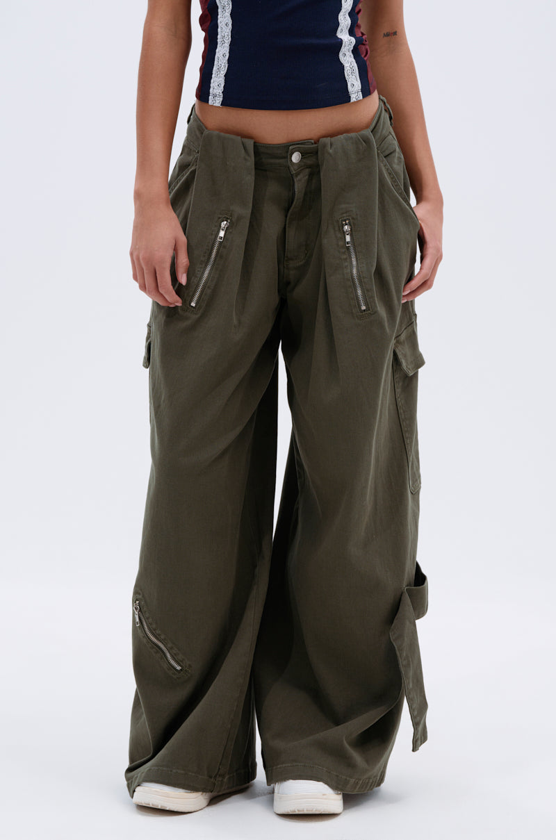 A person wears the COOL GIRL ERA TROUSER—oversized olive green cargo pants with a wide-leg silhouette and multiple zippered pockets—paired with a navy blue crop top with white lace trim. Only torso and legs are shown; background is plain white.