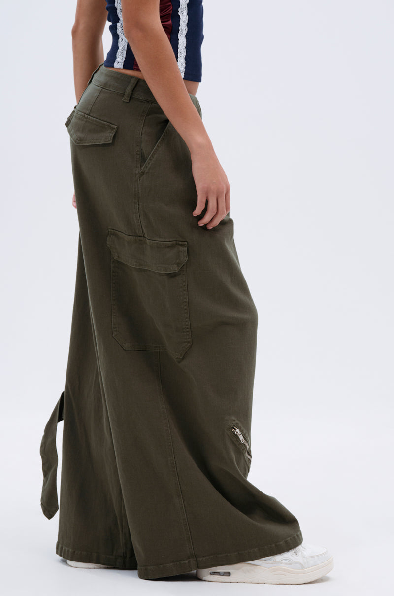 A person stands sideways against a plain light background wearing COOL GIRL ERA TROUSER cargo pants featuring a high-rise, wide-leg fit, and large pockets, paired with white sneakers.