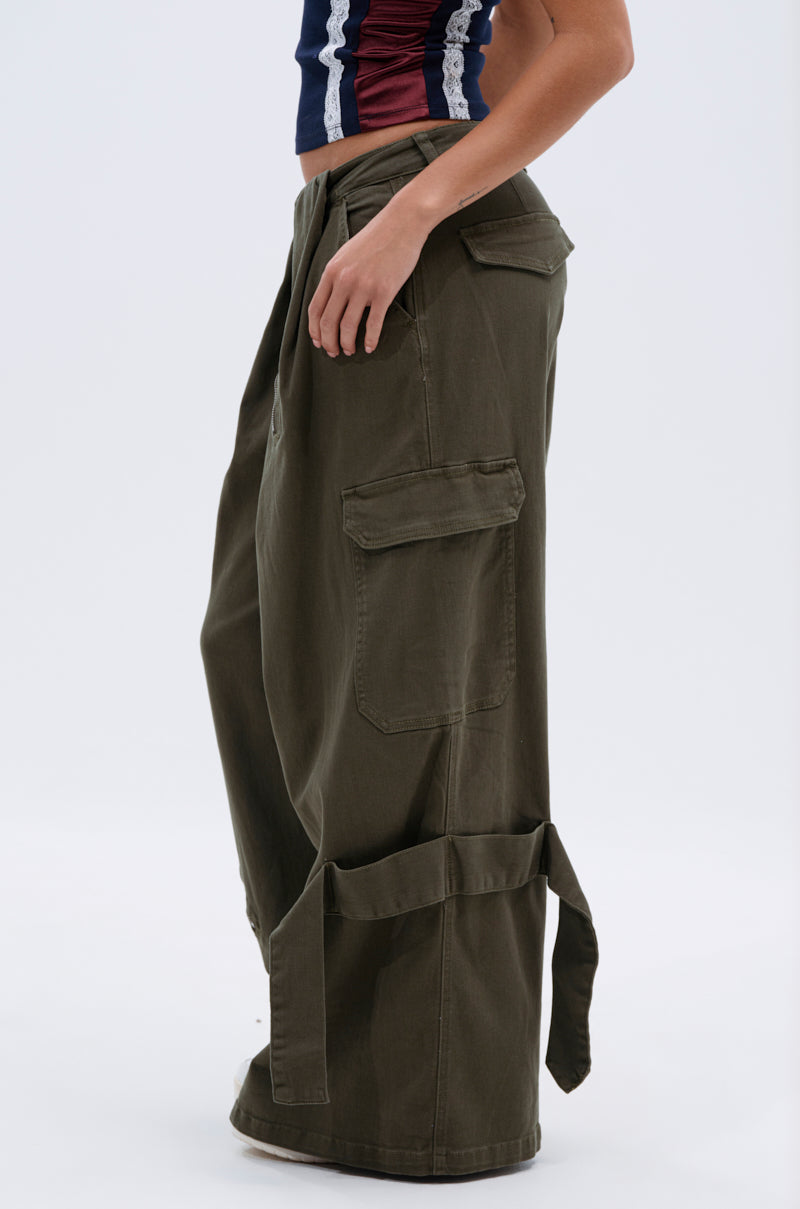 A person wearing the COOL GIRL ERA TROUSER—olive green, wide-leg cargo pants with large pockets and straps—paired with a sleeveless crop top in red, white, and blue vertical stripes. The background is plain and light-colored.