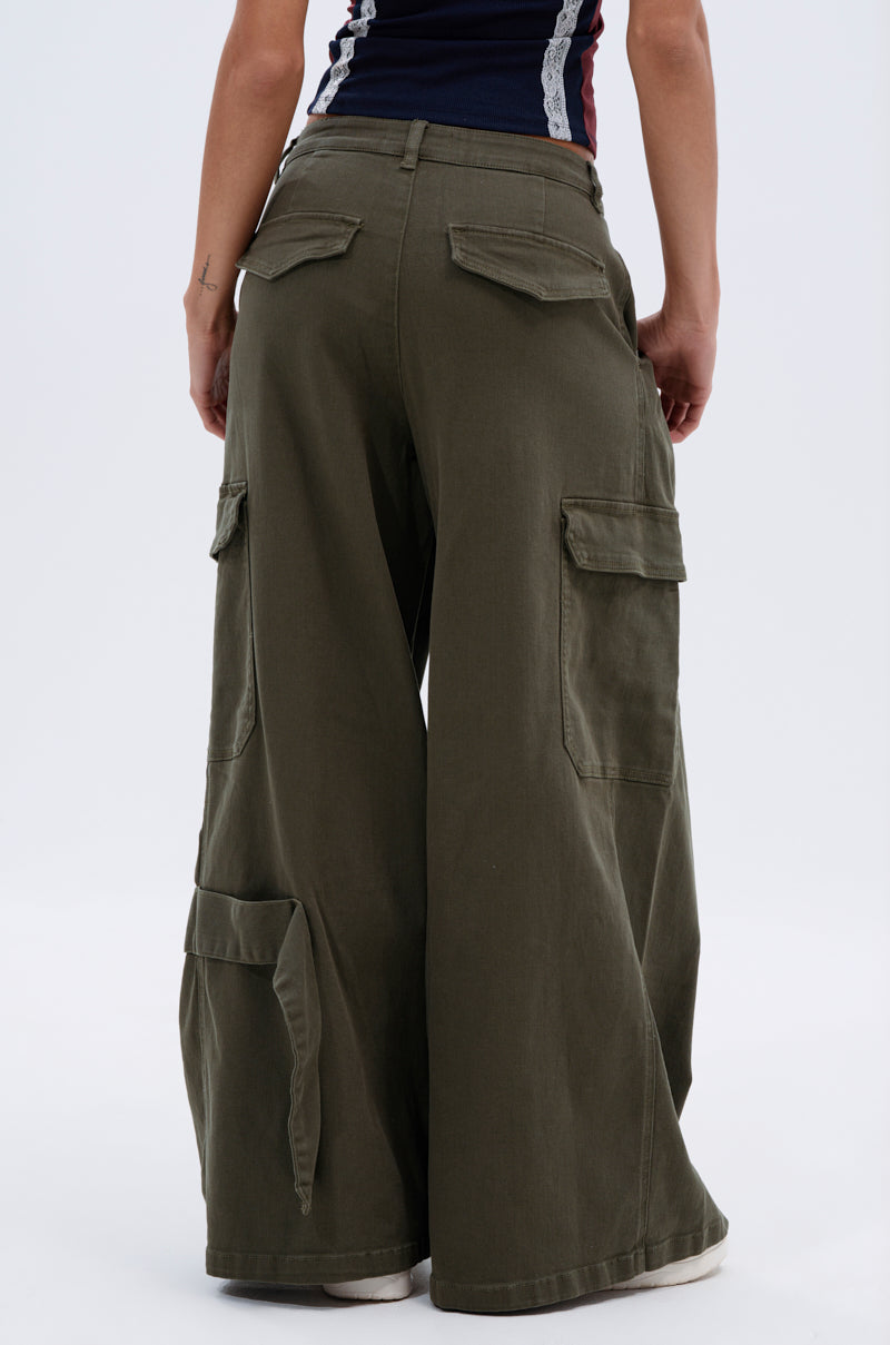 A person is seen from the back wearing the COOL GIRL ERA TROUSER—olive green cargo pants with a wide-leg fit and large pockets—paired with a dark sleeveless top and white shoes for a stylish, casual look.