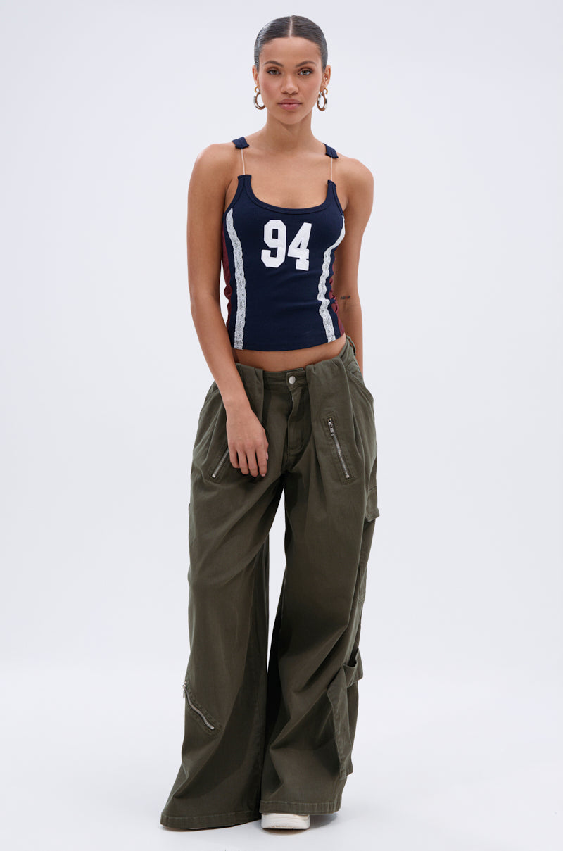 A woman stands against a plain background wearing a navy blue tank top with "94" on the front and COOL GIRL ERA TROUSER—olive green cargo pants with a wide-leg silhouette and zipper details.