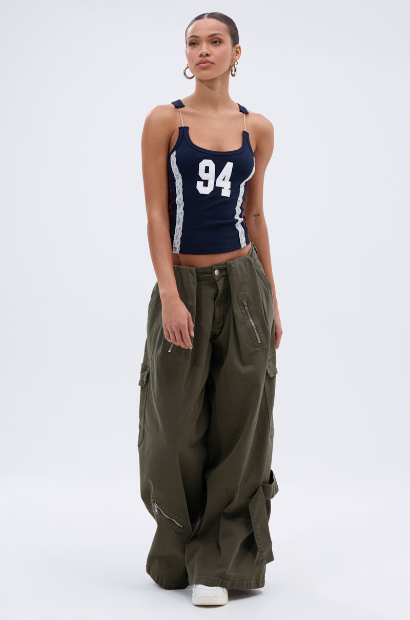 A woman poses against a plain background in a navy tank top with the number 94 and side stripes, COOL GIRL ERA TROUSER cargo pants in olive with a wide-leg silhouette and zippers, white sneakers, and hoop earrings.