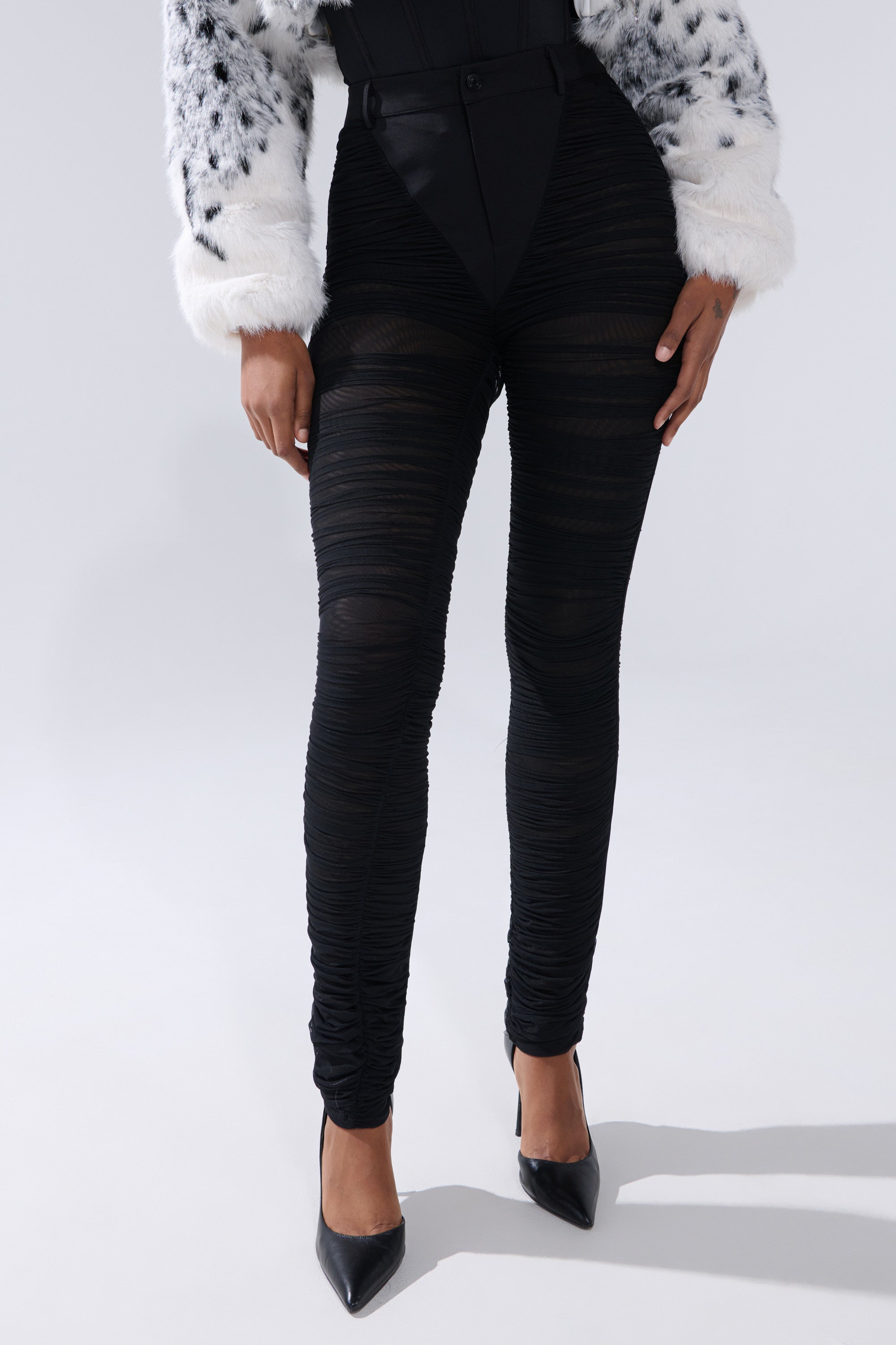 Wearing the TIGHT AND SEXY RUCHED TROUSER in black, a white faux fur jacket with black spots, and black pointed-toe heels, the person stands against a plain light background. The photo is chest-up cropped. 0
