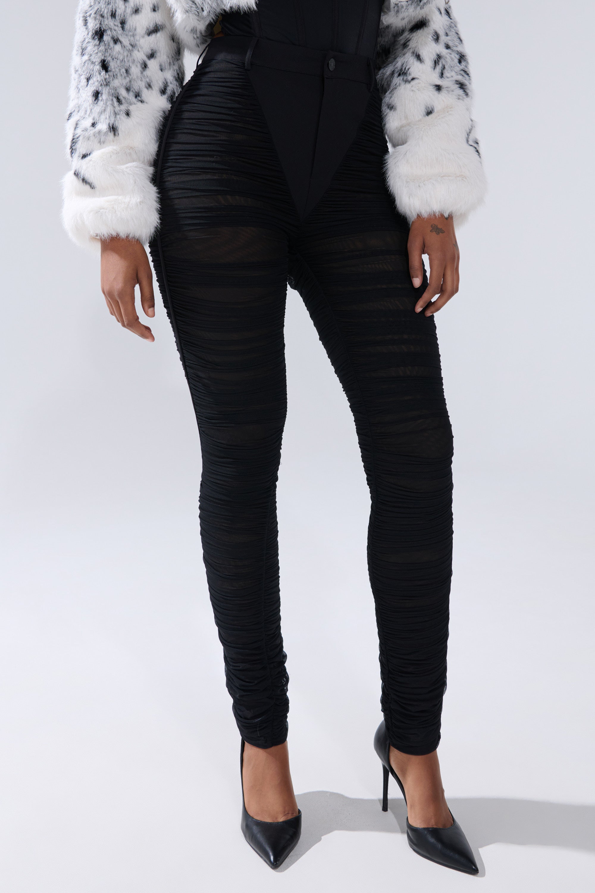 Someone stands against a plain background wearing the TIGHT AND SEXY RUCHED TROUSER, a pair of black sheer mesh high-rise pants, paired with black pointed-toe heels, a white faux-fur jacket with black spots, and a structured black top. 1