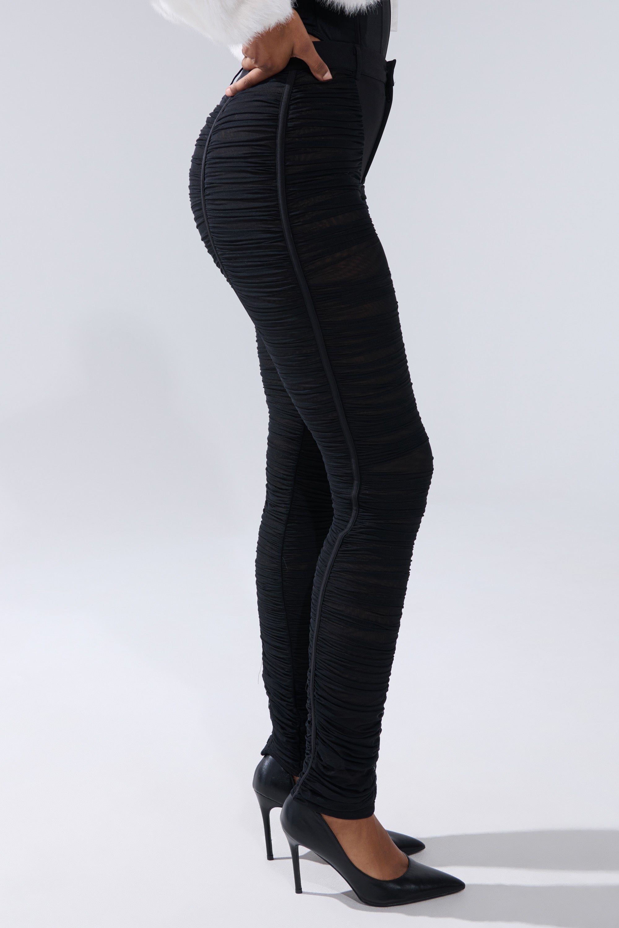 A person in the lower half is shown in profile against a plain light gray background, wearing black TIGHT AND SEXY RUCHED TROUSER paired with black high-heeled shoes. 2