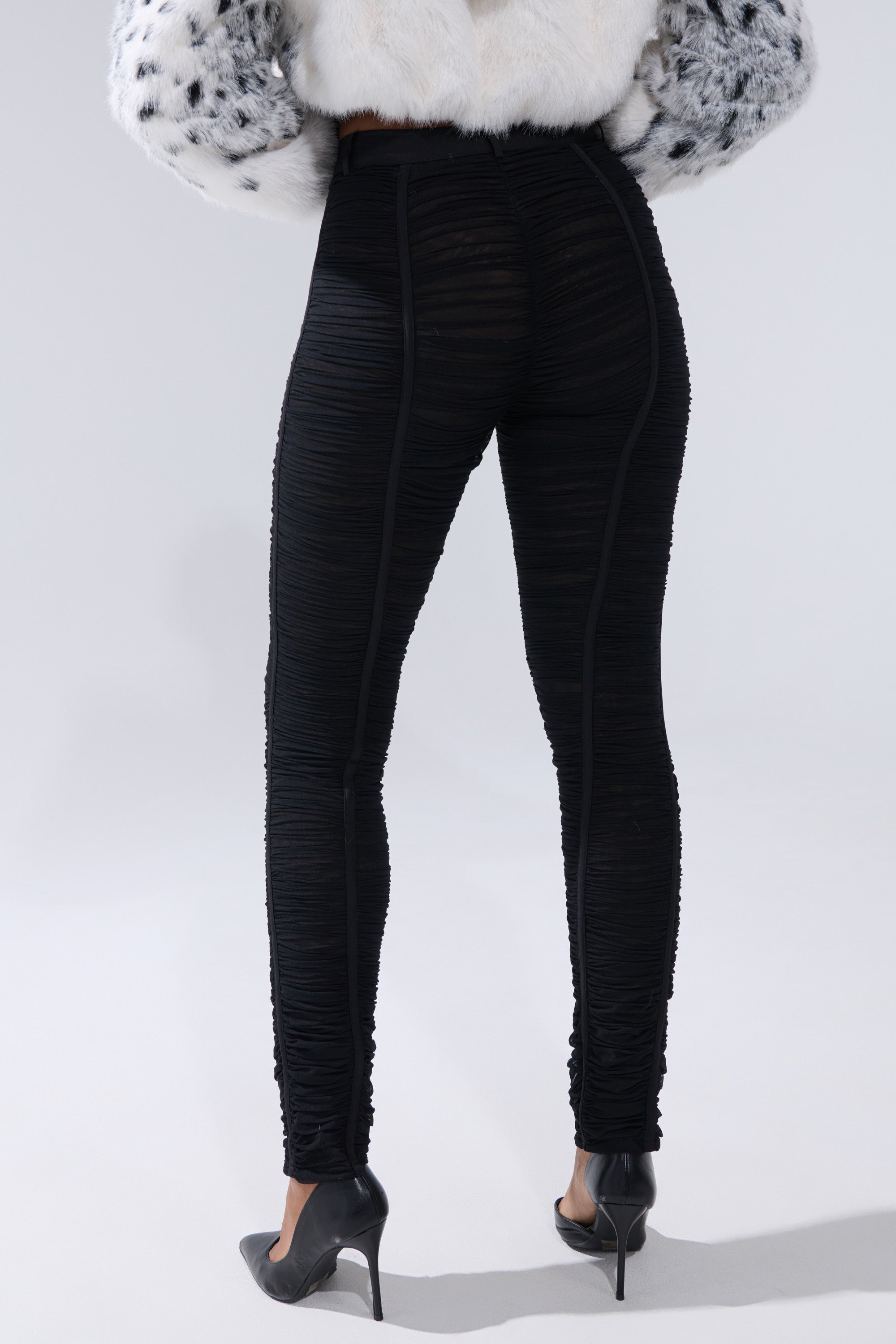 A person faces away, wearing the TIGHT AND SEXY RUCHED TROUSER with black high heels and a white faux fur coat with black spots, set against a plain light background. 3