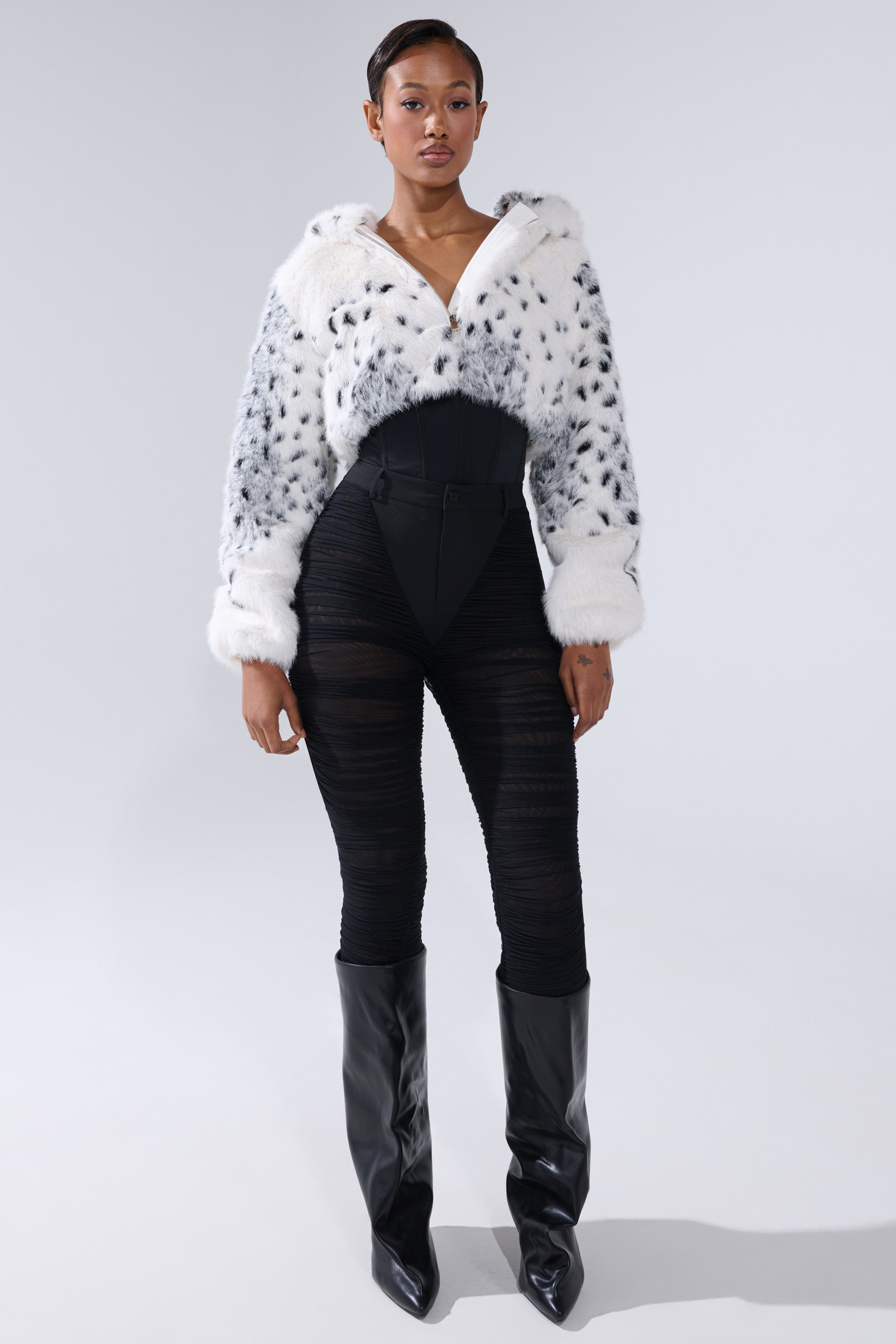 A woman faces forward in a white and black spotted faux fur jacket, TIGHT AND SEXY RUCHED TROUSER, and knee-high black boots against a plain light background. 4