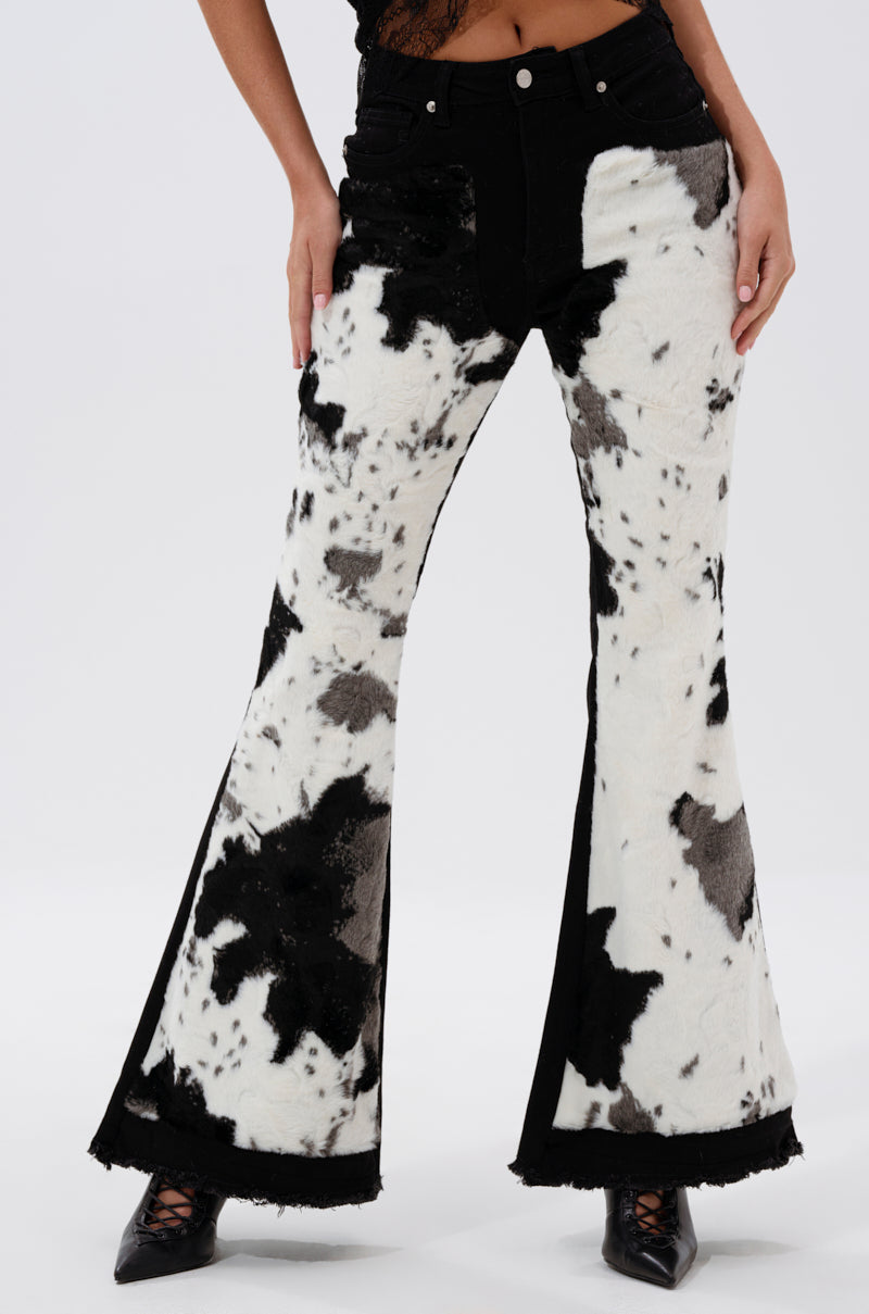 A person wears LETS GO TO THE RODEO BABY COW DENIM pants with black and white faux cowhide flared panels and a fuzzy texture, paired with black pointed-toe shoes. The upper body is mostly out of frame.