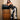 A woman with long dark hair in a black sequined top and pointed heels poses with a large black bag, wearing LETS GO TO THE RODEO BABY COW DENIM pants, leaning on a wooden barrel in a room with light walls and wood flooring.