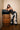 A woman with long dark hair in a black sequined top and pointed heels poses with a large black bag, wearing LETS GO TO THE RODEO BABY COW DENIM pants, leaning on a wooden barrel in a room with light walls and wood flooring.