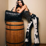 A woman with long dark hair in a black sequined top and pointed heels poses with a large black bag, wearing LETS GO TO THE RODEO BABY COW DENIM pants, leaning on a wooden barrel in a room with light walls and wood flooring.