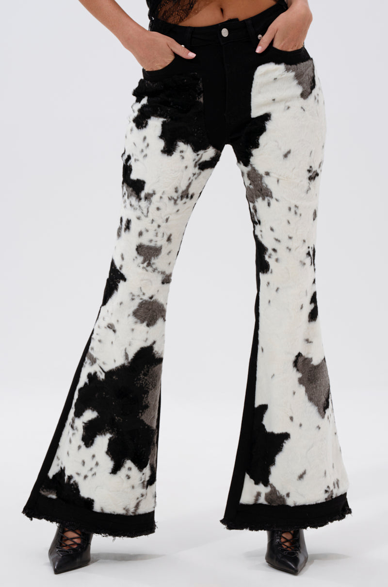 Wearing LETS GO TO THE RODEO BABY COW DENIM high-rise flare jeans with black pointed-toe heels, hands in pockets, against a plain white background for a modern take on western-inspired style.