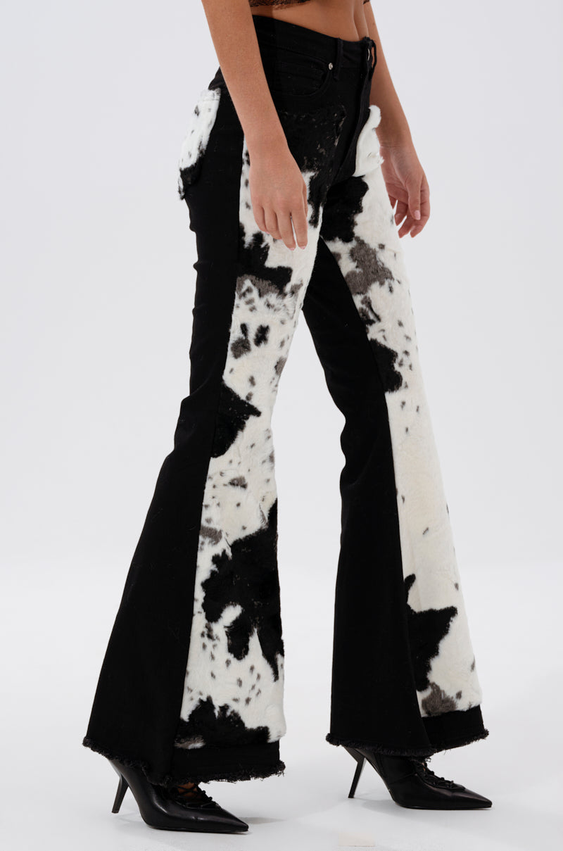 Someone wears LETS GO TO THE RODEO BABY COW DENIM—black high-rise flare jeans with white and black cow-print fuzzy panels—paired with black pointed-toe heeled shoes, set against a plain white background.