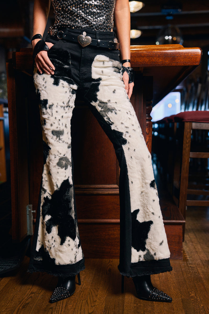A person stands indoors, showing only their lower half, wearing LETS GO TO THE RODEO BABY COW DENIM high-rise flare jeans, a sparkling sleeveless top, black spiked heels, and a belt with a heart-shaped buckle.