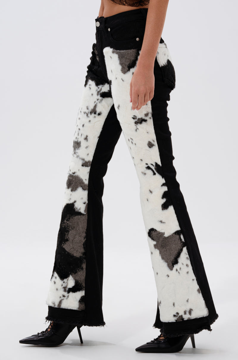 Wearing the LETS GO TO THE RODEO BABY COW DENIM—high-rise, cow print jeans with textured, fuzzy fabric—paired with black pointed-toe heels. Only the lower torso and legs appear against a plain background, highlighting the bold western style.
