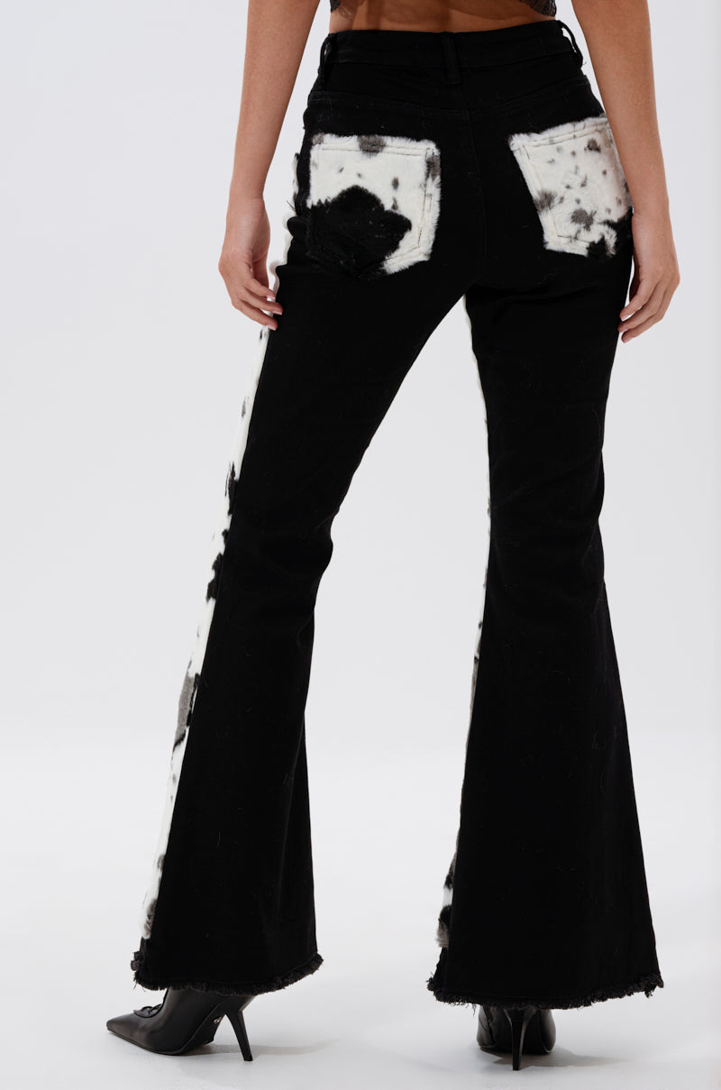 Wearing the LETS GO TO THE RODEO BABY COW DENIM, a person stands facing away, showing off the black high rise flare jeans with white and black cow print patches on the back pockets and sides. They pair them with black pointed-toe heels.