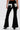 Wearing the LETS GO TO THE RODEO BABY COW DENIM, a person stands facing away, showing off the black high rise flare jeans with white and black cow print patches on the back pockets and sides. They pair them with black pointed-toe heels.