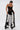 A woman styles the LETS GO TO THE RODEO BABY COW DENIM pants with a sheer black lace bustier top and black heels. Her slicked-back hair and bold choker necklace complete the look.