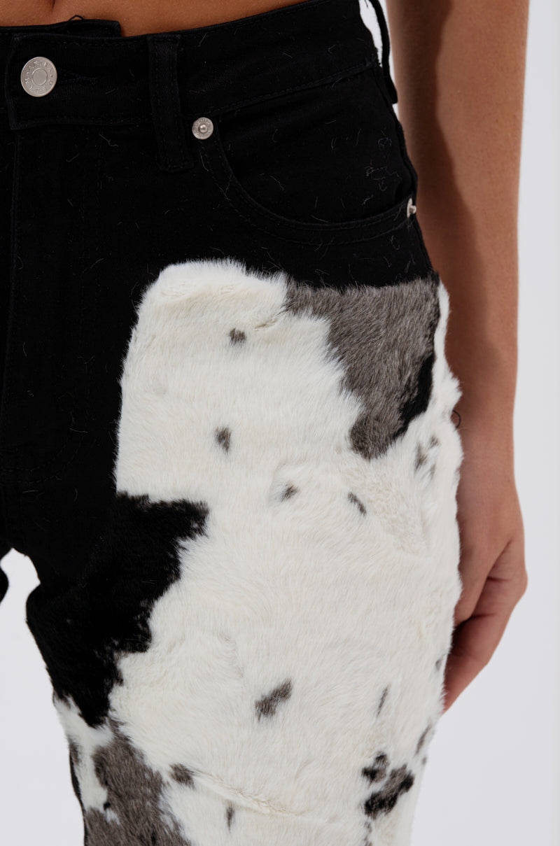 Close-up of a person's waist and hip in LETS GO TO THE RODEO BABY COW DENIM high-rise flare jeans with a cow print and white-gray faux fur patch on the upper thigh; their arm rests at their side.