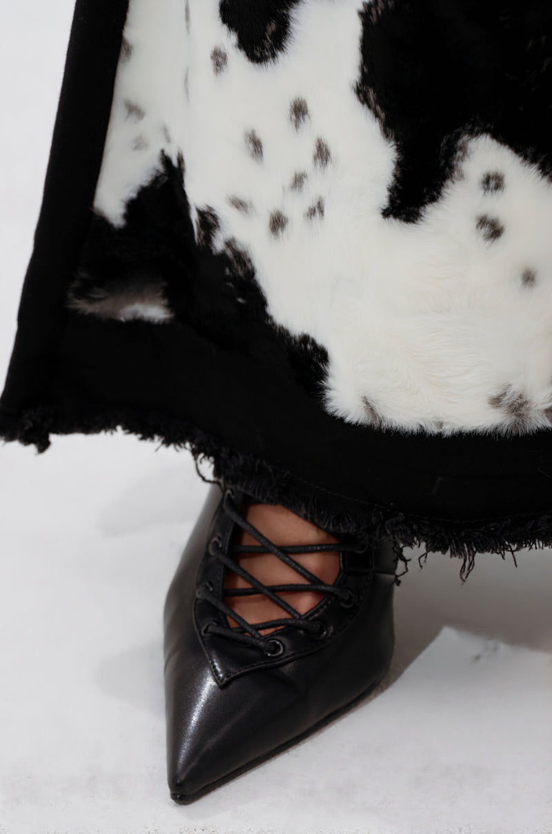 A close-up shows a person wearing pointed black lace-up shoes and the LETS GO TO THE RODEO BABY COW DENIM, with its cow print hem visible above the shoes against a white background.