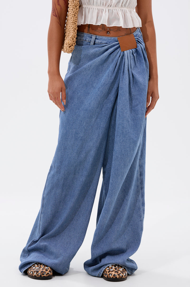 Someone wearing the TOO MUCH FOR YOU WIDE LEG DENIM with draped details, a brown belt, white sleeveless top, woven bag, and leopard-print shoes stands against a plain light background.