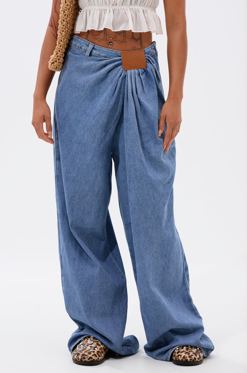 Wearing TOO MUCH FOR YOU WIDE LEG DENIM, a white ruffled crop top, tan belt, woven bag, and leopard-print shoes, a person stands against a plain white background.