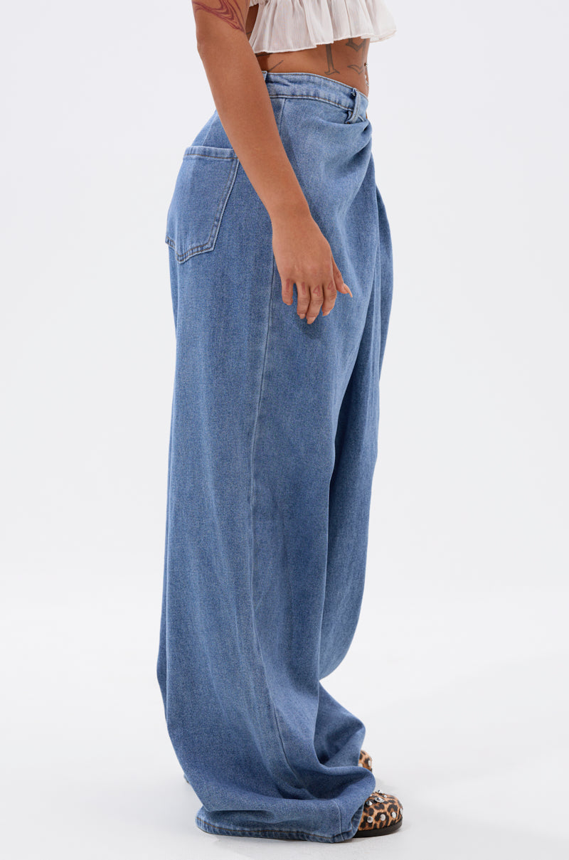 A person stands sideways in TOO MUCH FOR YOU WIDE LEG DENIM jeans and a white cropped top, showing a left arm tattoo and wearing leopard print shoes against a plain white background.