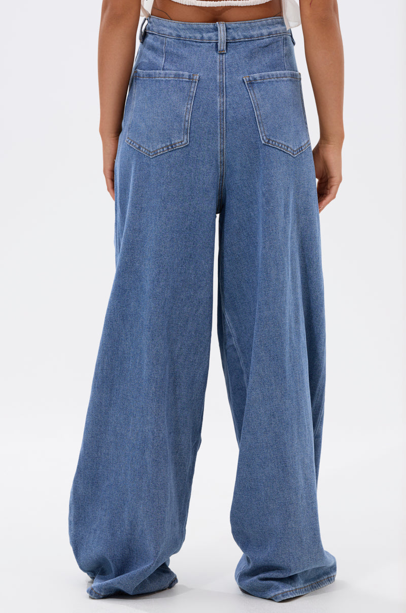 A person wearing the TOO MUCH FOR YOU WIDE LEG DENIM—high-waisted, loose-fitting blue jeans with large back pockets, pleated front, and floor-length hems—is seen from behind, paired with a white top.