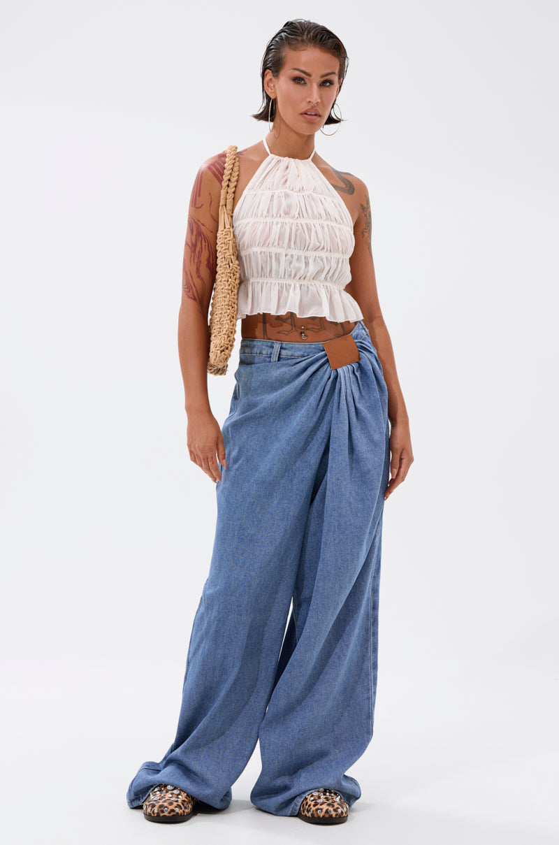 A woman poses confidently in a sleeveless white ruffled crop top, TOO MUCH FOR YOU WIDE LEG DENIM jeans with pleats, animal print shoes, and a woven shoulder bag. Her wet-look hair and arm tattoos stand out against a white background.
