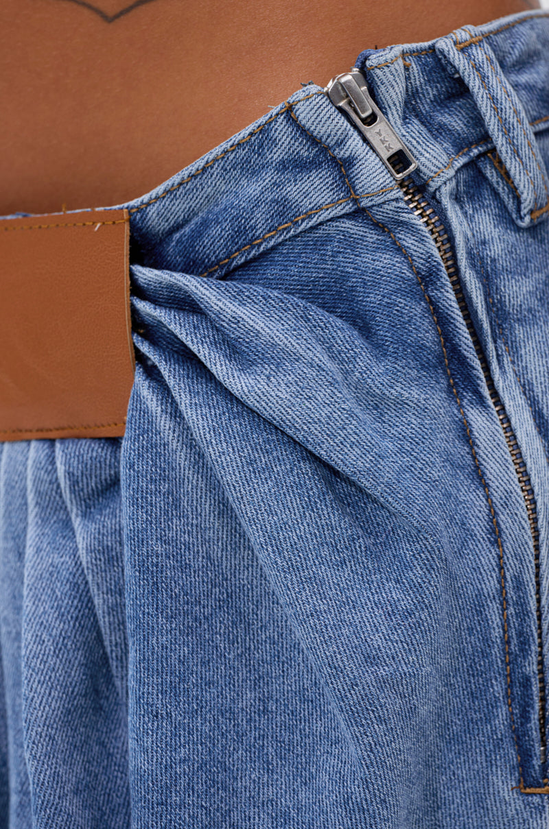 Close-up of the TOO MUCH FOR YOU WIDE LEG DENIM jeans in blue, highlighting the high-rise, pleated front near the waistband, brown leather patch detail, and unzipped metal zipper. A hint of waist and skin appears at the top.