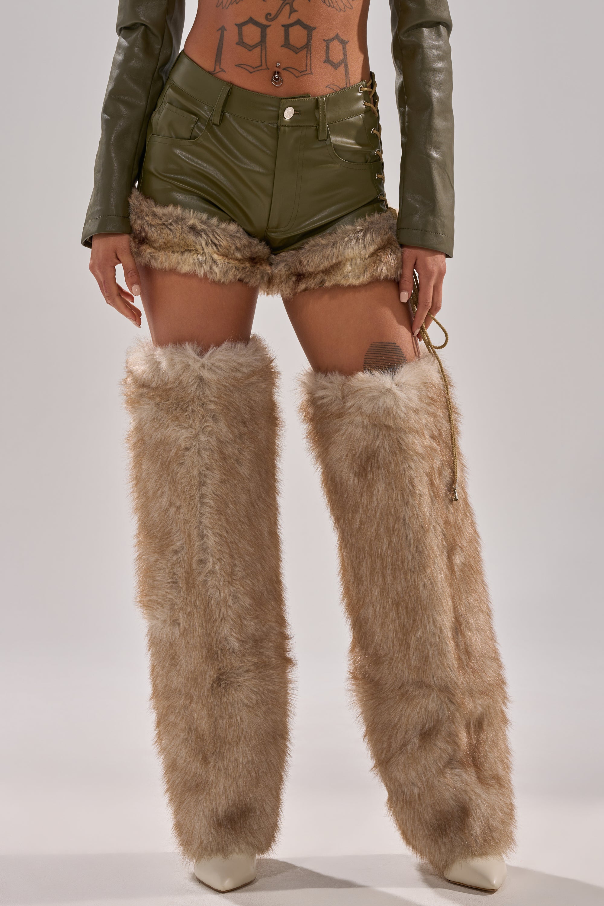 Someone wearing TRAMP STAMP FAUX LEATHER SHORTS in olive green with faux fur trim, paired with tan furry leg warmers over white boots and a cropped top revealing a "1999" tattoo design. The backdrop is plain and neutral.