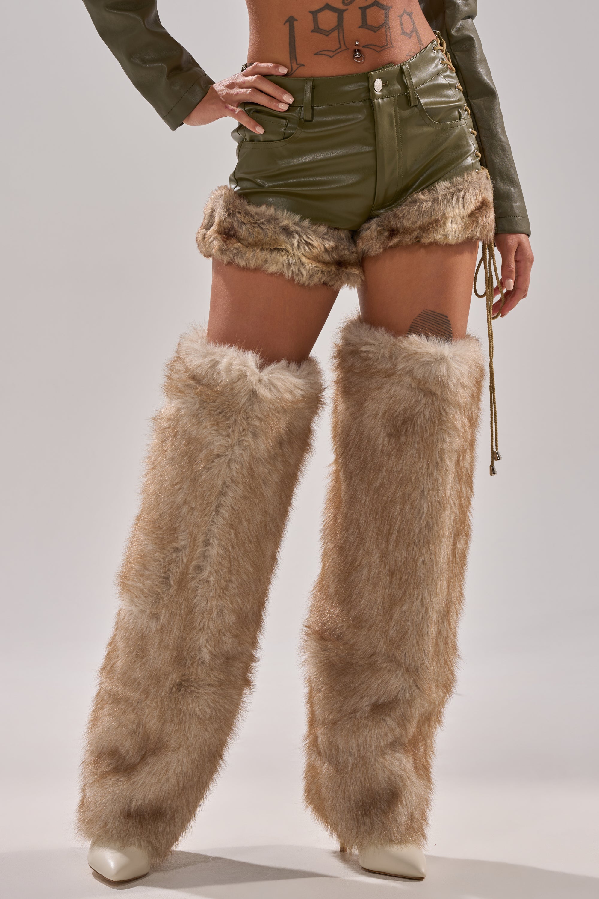 Someone wearing TRAMP STAMP FAUX LEATHER SHORTS, beige furry leg warmers, white boots, and showing a “1999” tramp stamp tattoo on their lower abdomen.