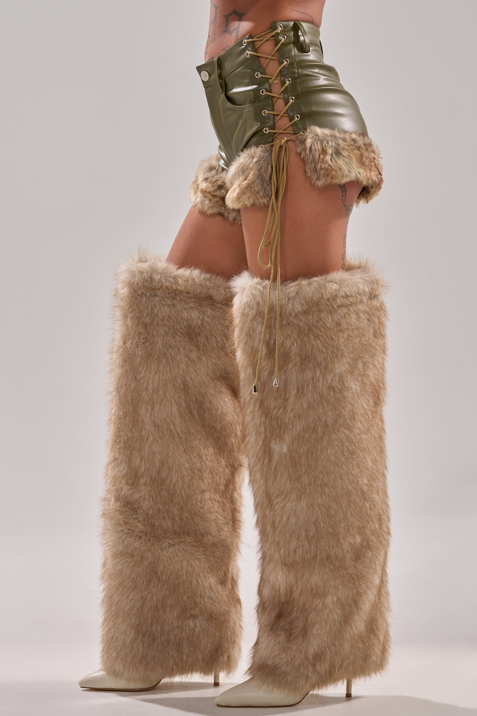 Wearing AKIRA Label's TRAMP STAMP FAUX LEATHER SHORT, a person pairs them with knee-high, tan faux fur stiletto boots and stands against a light background.