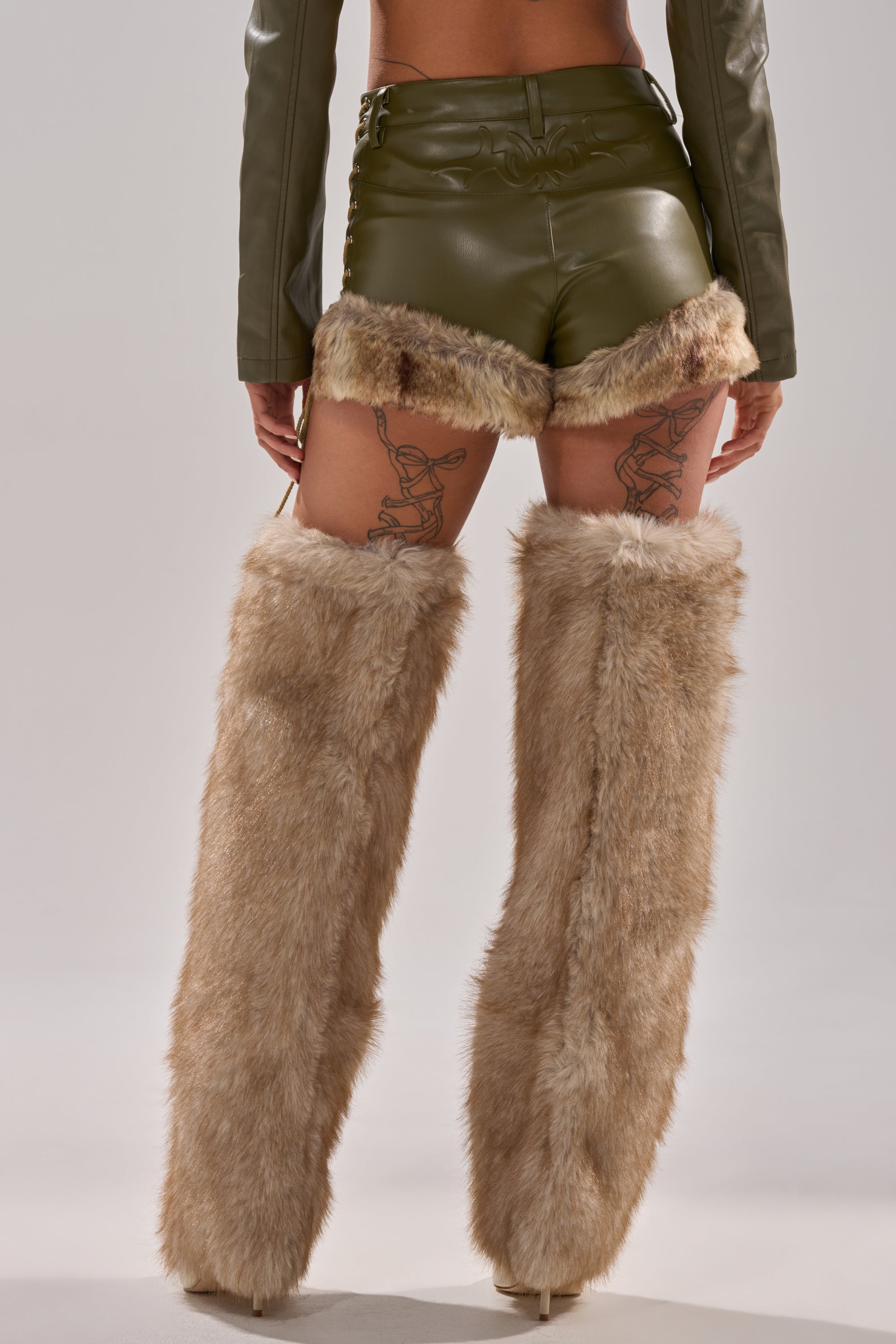 A person stands facing away, wearing green TRAMP STAMP FAUX LEATHER SHORTS with fur trim, a matching long-sleeve top, and tan faux fur leg warmers over high heels. A tattoo design is visible on the back of their thighs.