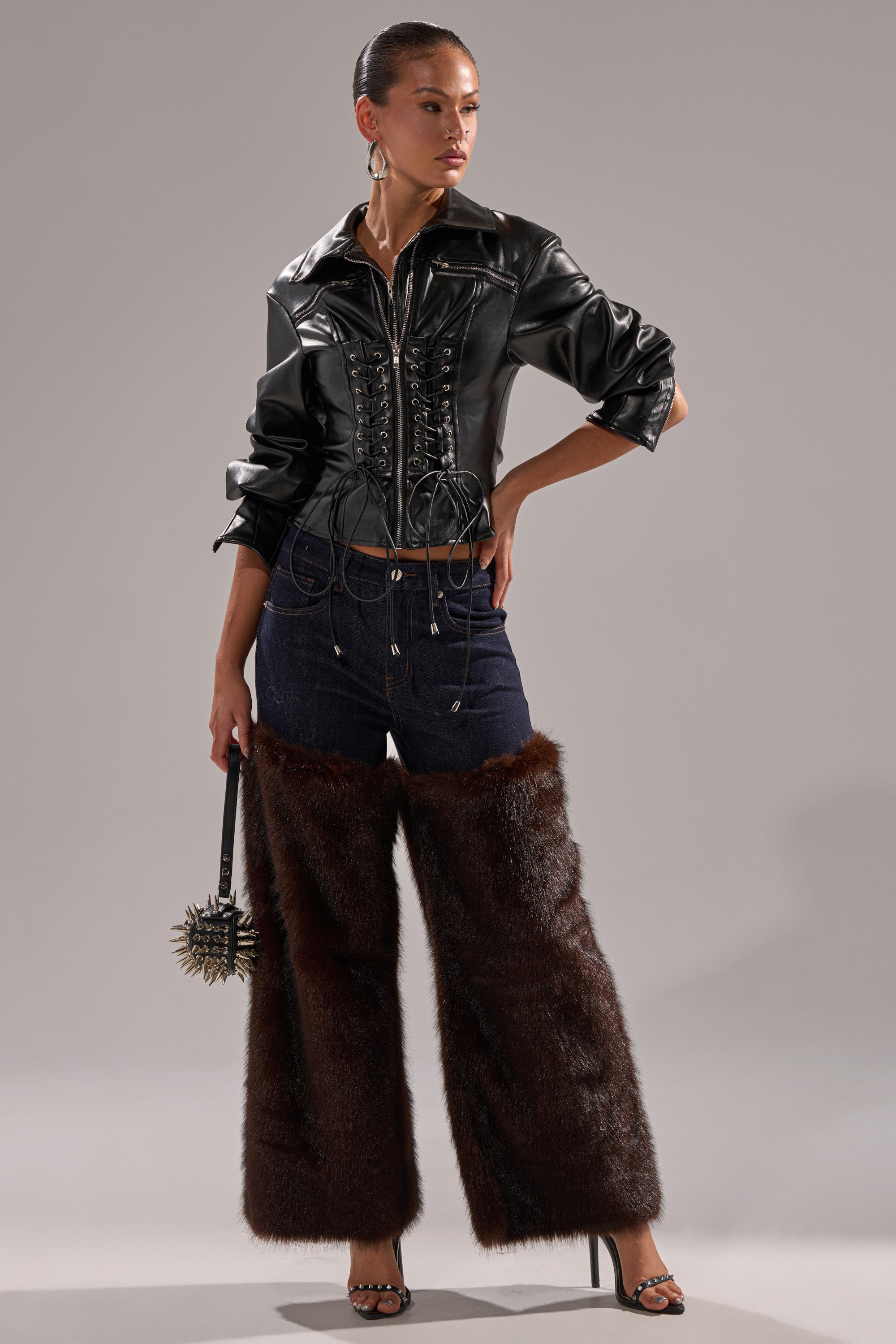 A model poses in the BOOTS WITH THE FUR WIDE LEG DENIM, a black leather corset jacket, black high-heeled sandals, hoop earrings, and holds a spiked clutch against a gray backdrop.