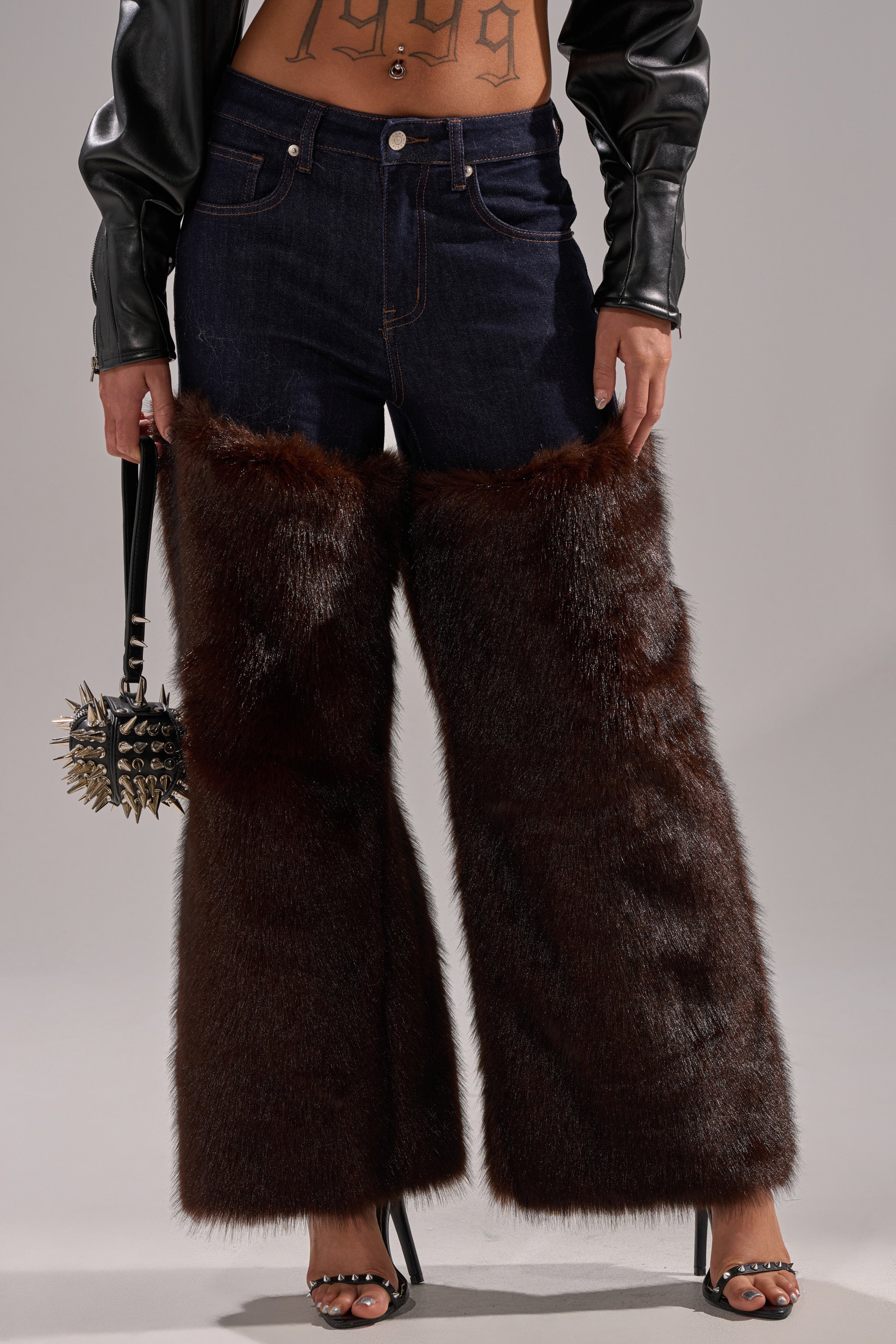 A person wears BOOTS WITH THE FUR WIDE LEG DENIM with large brown faux fur trim, a leather jacket, open-toe heels, and carries a black spiked handbag. A "1999" tattoo is visible on their torso.