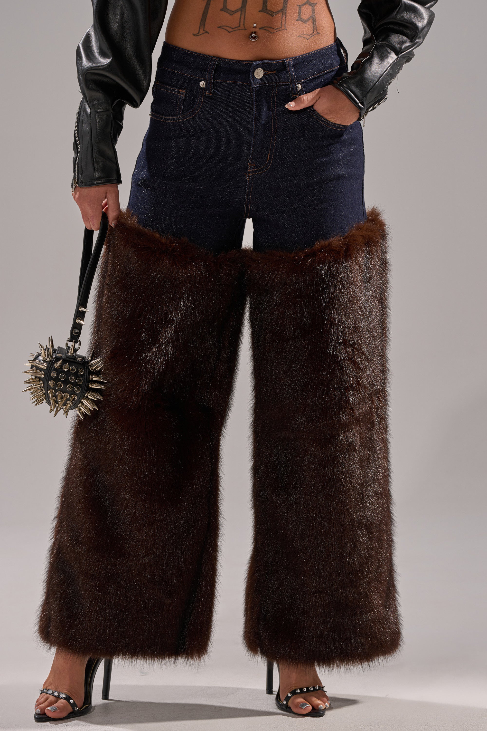 A person wears BOOTS WITH THE FUR WIDE LEG DENIM featuring oversized brown faux fur panels, styled with black open-toe heels, a black leather jacket, and a spiked handbag. Tattoos decorate their midriff.