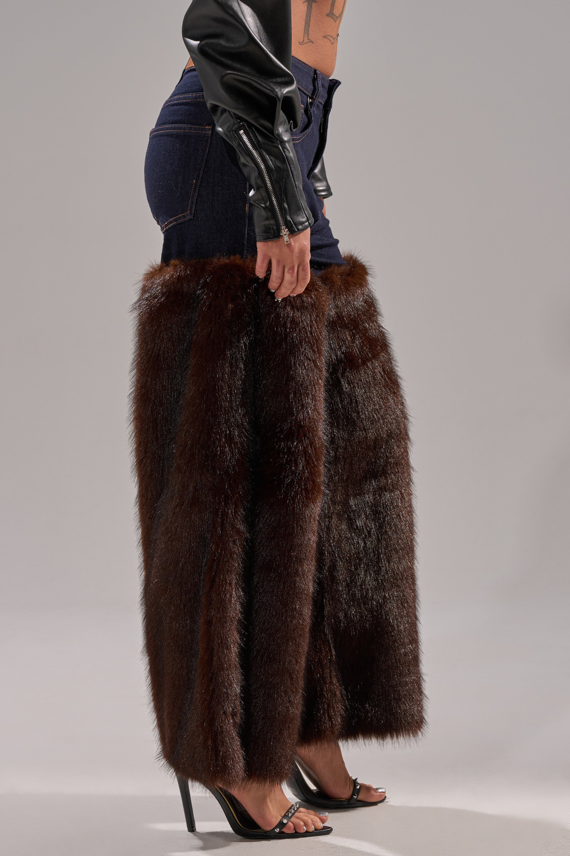 A person models the BOOTS WITH THE FUR WIDE LEG DENIM—high-rise jeans with floor-length brown faux fur chaps—styled with a black leather jacket and open-toe heels, standing before a plain gray backdrop.