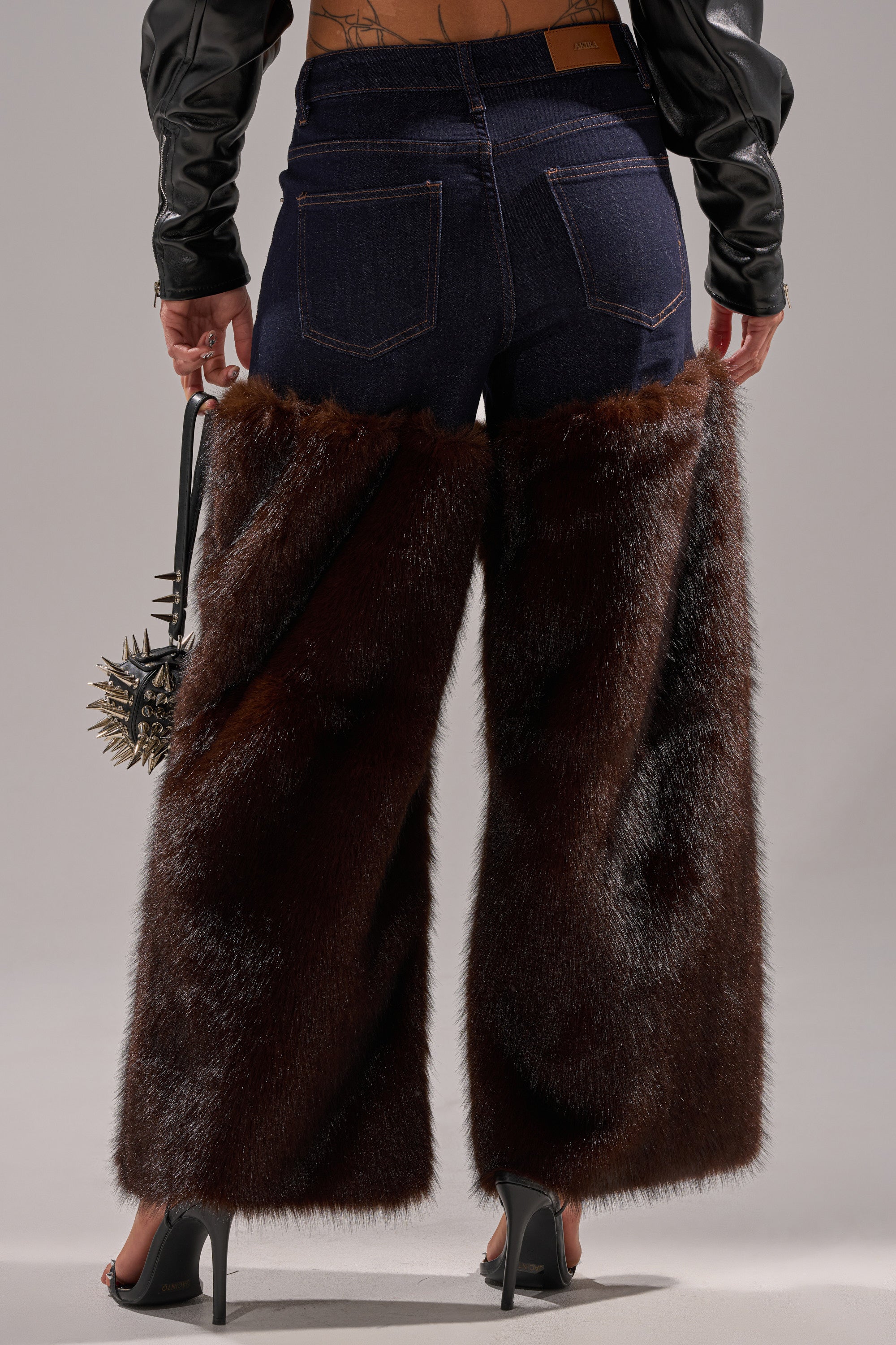 A person faces away, wearing BOOTS WITH THE FUR WIDE LEG DENIM—high-waisted jeans with furry chaps—plus a spiked handbag, black high heels, and a cropped top with black leather sleeves.